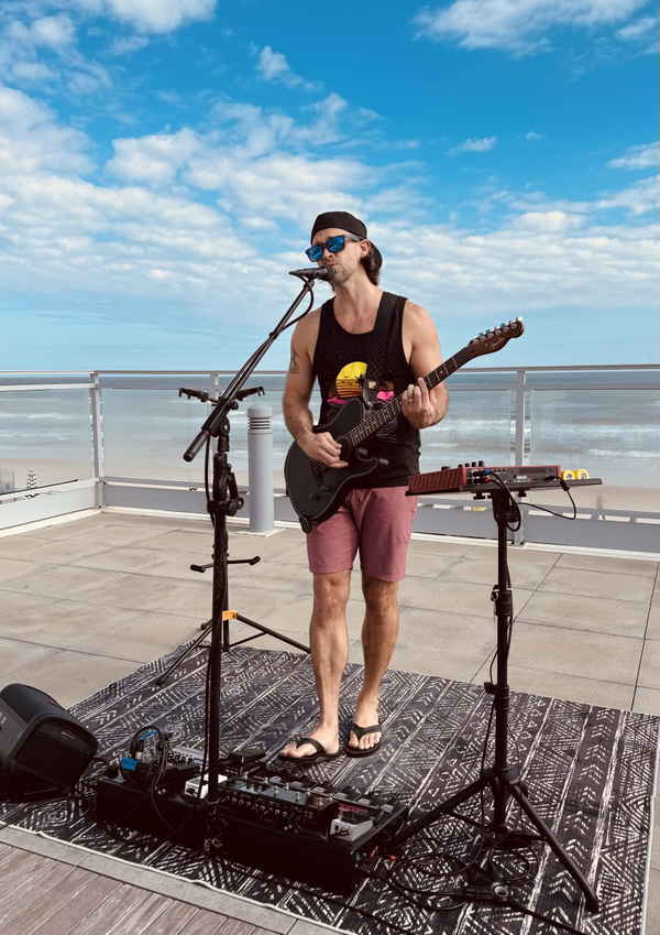 Our male loop music artist happily performing on patio by beach shore while wearing shorts tank top and backwards cap | Altus Entertainment
